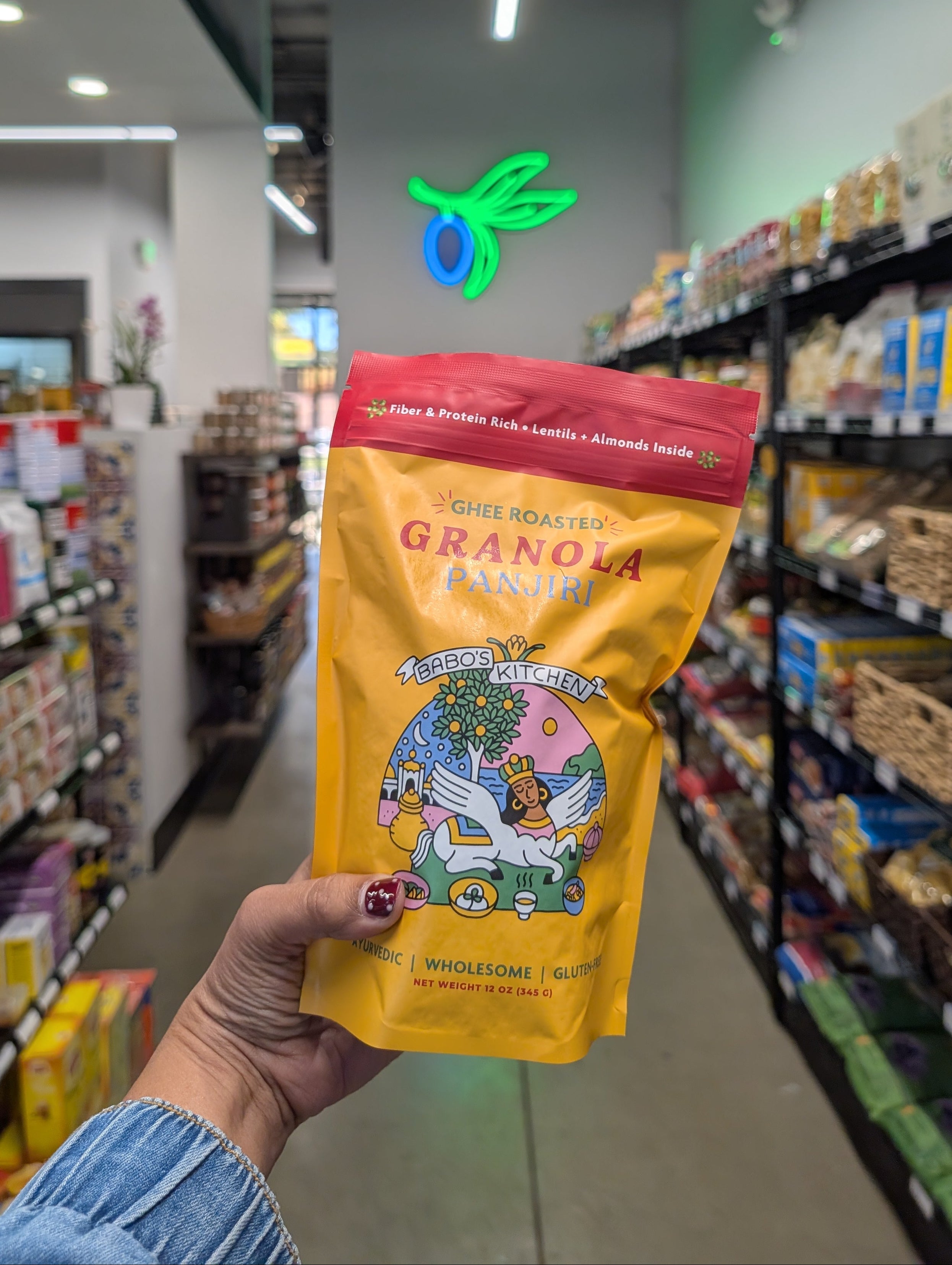 A hand holds Babo's Panjiri Granola in a grocery store aisle, with shelves of products blurred in the background.